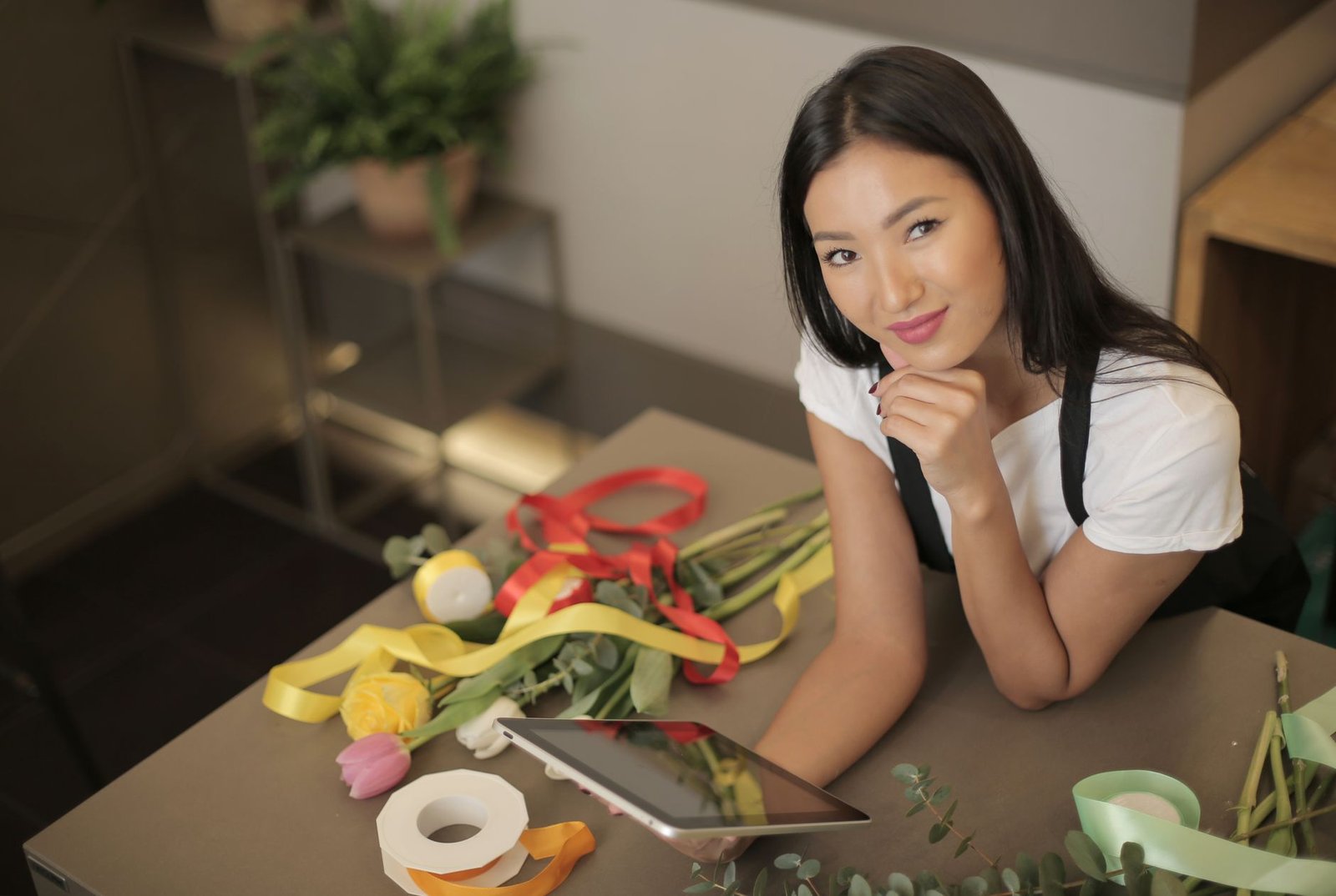 A young florist in a flower shop smiles while arranging flowers and using a tablet.